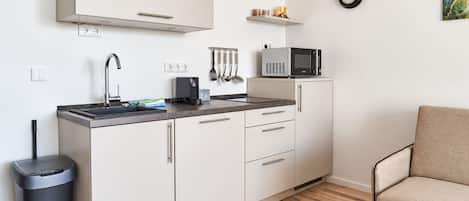 A compact kitchenette with a microwave, kettle, and utensils on the counter, a trash bin, and a chair in the corner.