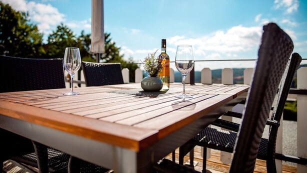 A wooden table with two wine glasses and a bottle of wine, set on an outdoor patio with wicker chairs.