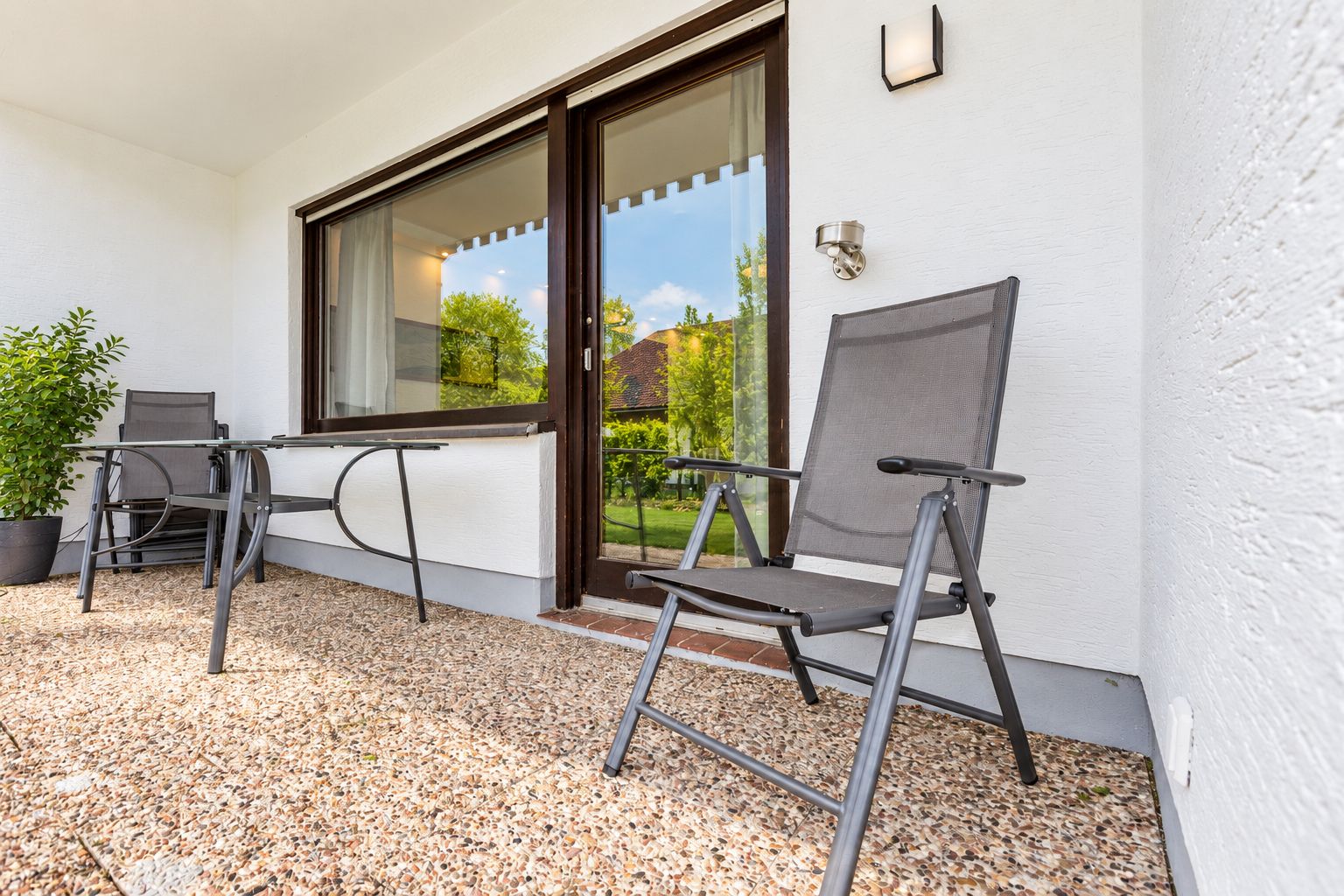 A patio with two chairs and a table, a potted plant, and a view of a house through a glass door.