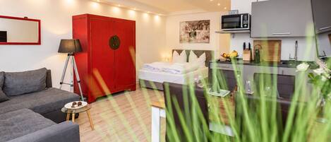 A modern hotel room with a bed, a red wardrobe, a kitchenette, and a grey sofa.