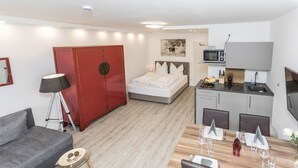 Ein modernes Hotelzimmer mit einem Bett, einem Esstisch für zwei Personen, einer Küchenzeile, einem roten Kleiderschrank und einem grauen Sofa.