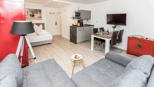 Ein modernes Wohnzimmer mit grauen Sofas, einem kleinen runden Tisch mit Snacks, einem Bett mit weißen Bettwäsche, einem roten Schrank und einer Küchenzeile mit Mikrowelle und Fernseher.