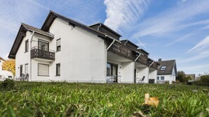 Ein modernes, zweistöckiges Haus mit weißer Fassade, einem Balkon im Obergeschoss und einer Rasenfläche im Vorgarten.