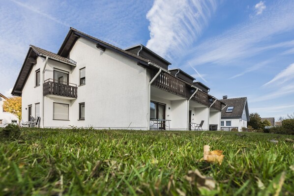 Une maison moderne à deux étages, avec une façade blanche, un balcon à l’étage supérieur et une pelouse devant.