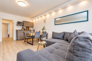 A modern living room with a grey sofa, a dining table with chairs, a kitchen area, and a large mirror on the wall.