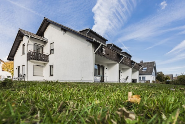 A modern two-story house with a white exterior, a balcony on the upper floor, and a grassy front yard.