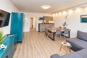 A modern living room with a grey sofa, a dining area with a wooden table and chairs, a kitchen with a microwave, and a teal-colored cabinet.