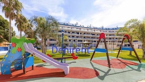 Un terrain de jeux avec des toboggans et des balançoires, entouré de palmiers et, au fond, un bâtiment doté de balcons.