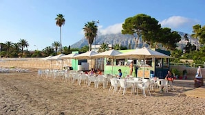 Un restaurant situé en bord de mer, avec une terrasse, offrant une vue sur les montagnes et bordé de palmiers.