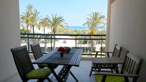 Un balcon avec une table et des chaises, donnant sur une plage bordée de palmiers et où l’on aperçoit une voiture rouge sur la route.