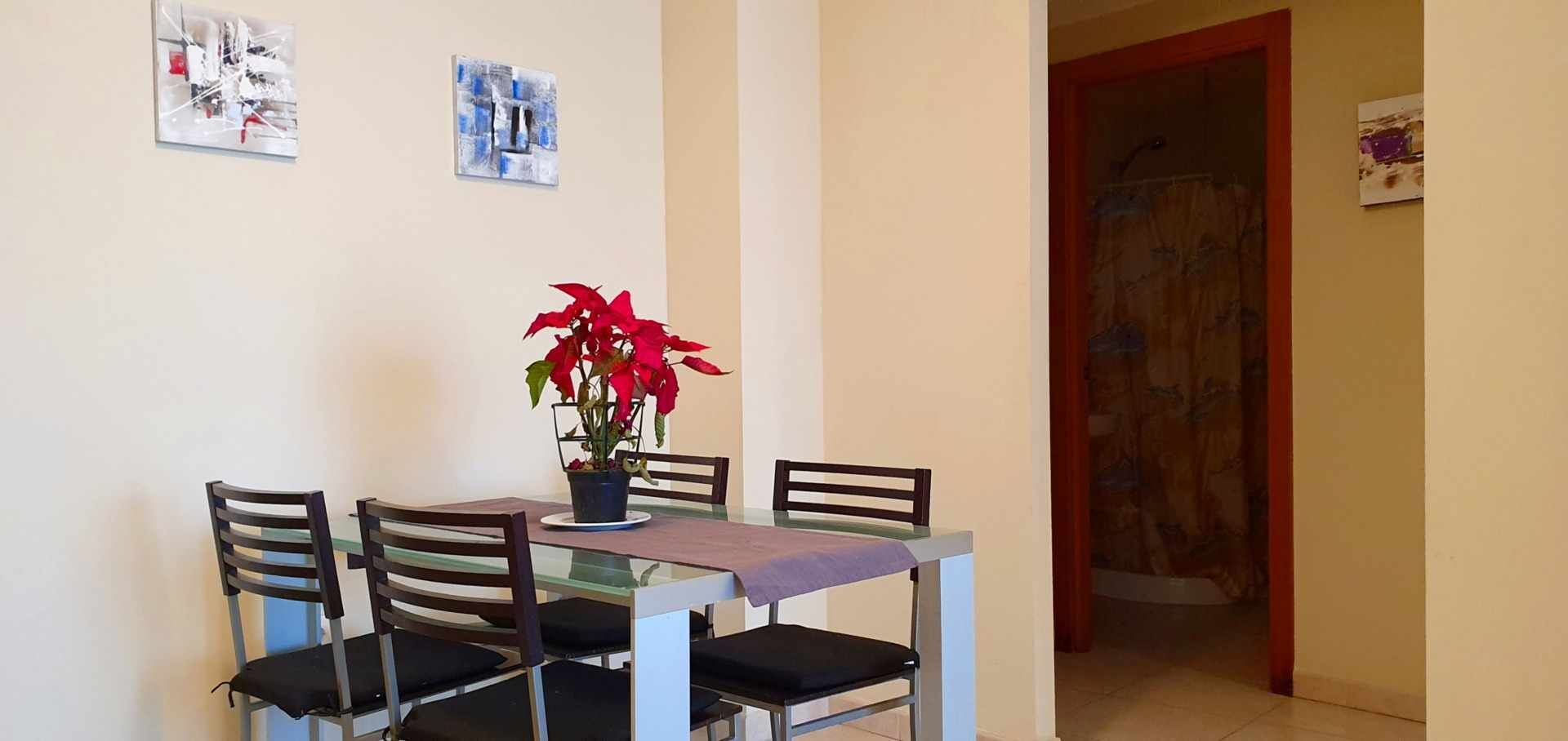 Un coin repas avec une table en verre, quatre chaises et une plante en pot ornée de fleurs rouges.