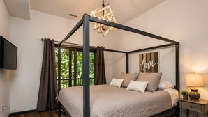 Ein Bett mit Baldachin, mit einer beigen Steudecke, ein Nachttisch mit Lampe und ein an der Wand befestigtes Fernsehgerät.