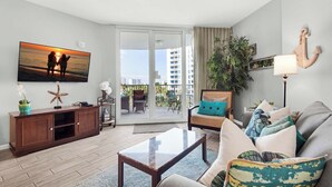 A living room with a large TV, a sofa, a coffee table, and a sliding glass door leading to an outdoor patio. - 🌴Beach Nearby+ Lagoon Pool -Palms 1505☀️ (Destin)