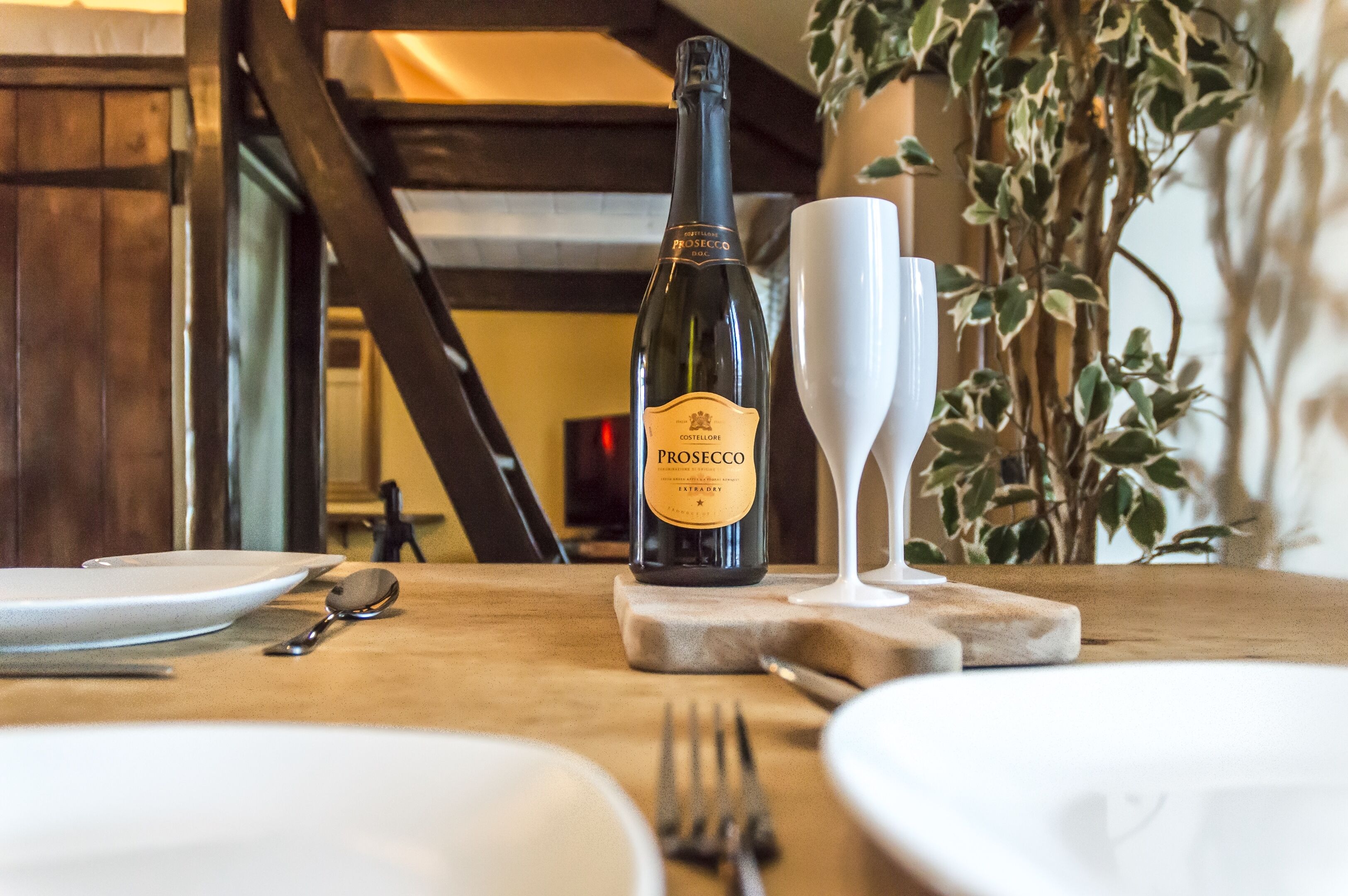 Una botella de Prosecco sobre una mesa de madera, con un plato blanco, una cuchara y un tenedor.