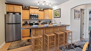 Eine moderne Küche mit einem Edelstahlkühlschrank, einer Mikrowelle und einer Kaffeemaschine. Holzschränke und eine Bar mit Barhocker sind ebenfalls vorhanden.