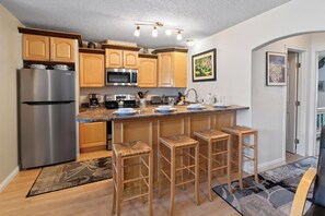 A modern kitchen with a stainless steel refrigerator, a microwave, and a coffee maker. There are wooden cabinets and a bar stool area. - Right on Main Street Amazing Views of the Ski Area (Breckenridge)