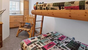 Ein Etagenbett mit Steppdecke, ein Holzstuhl mit Kissen, ein Schreibtisch und ein Fenster mit Jalousien.