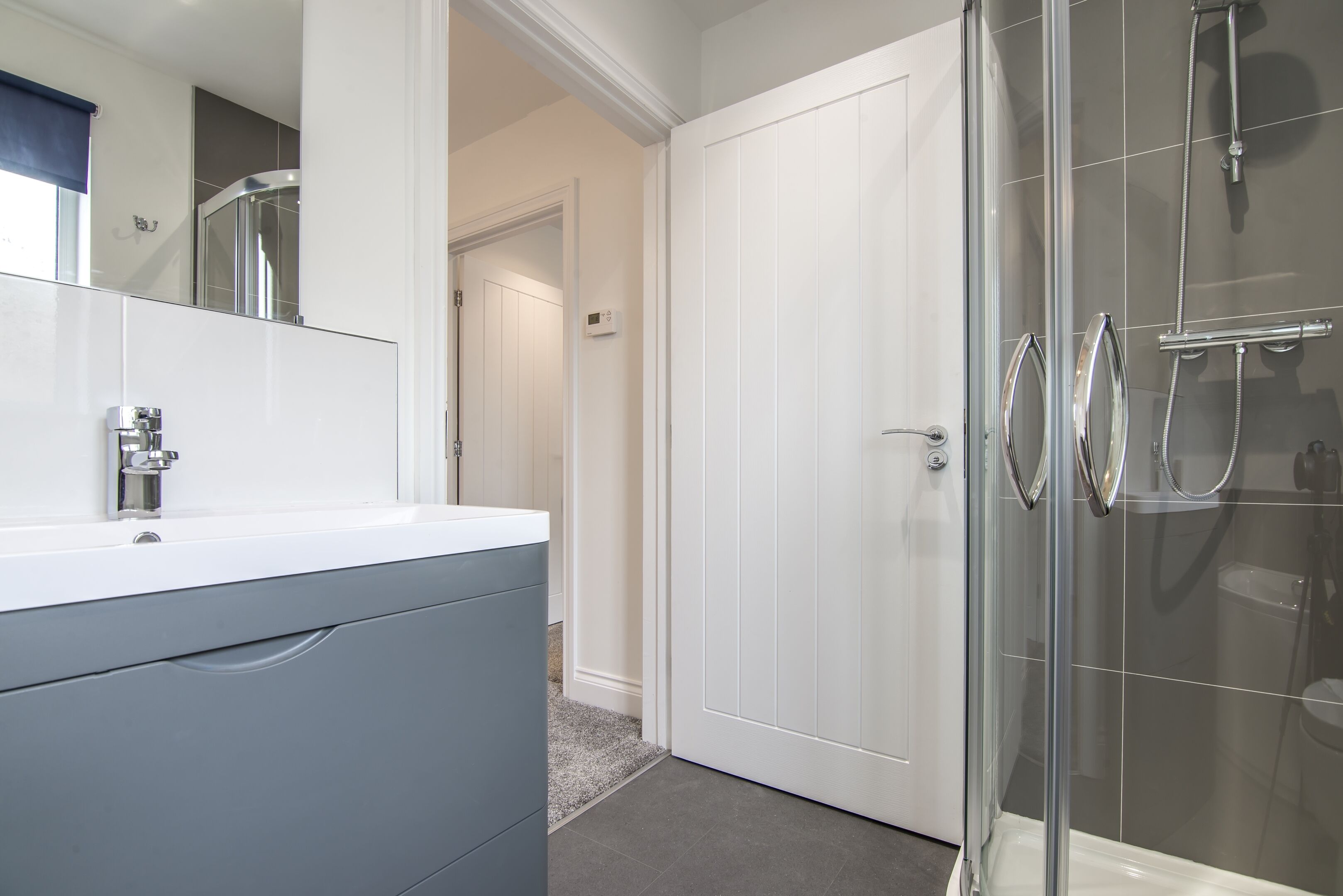 A bathroom with a glass shower enclosure, a white vanity with a mirror, and a door leading to another room.