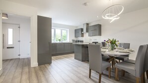 A modern kitchen with grey cabinets, a central island, and a dining area with a round table and chairs. - Martello View - 3 Bedroom - Llanreath (Pembroke Dock)