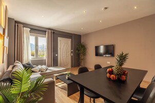 Una sala de estar moderna con un sofá, una mesa de comedor con fruta, un televisor y un amplio ventanal con cortinas.