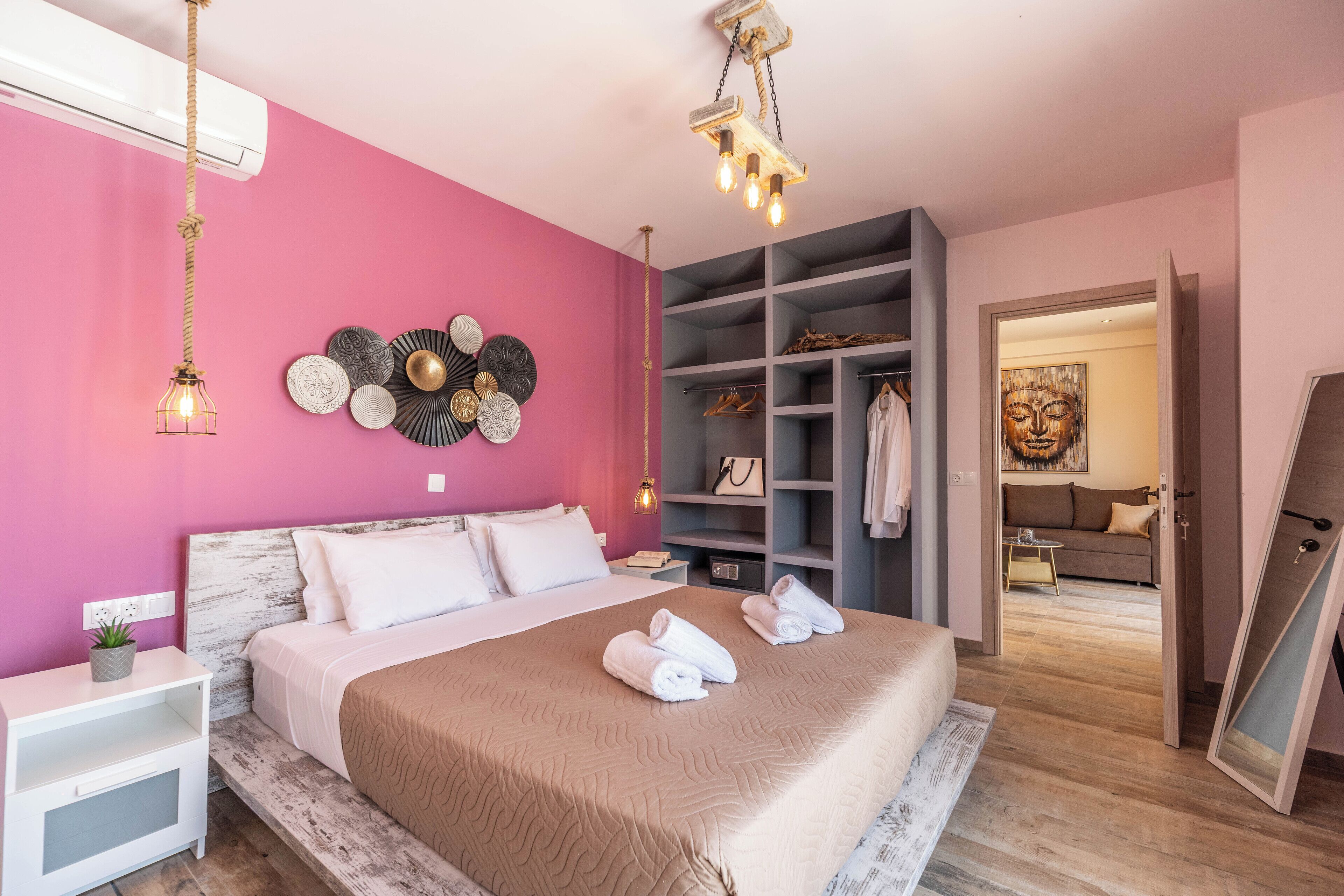 Een slaapkamer met een roze muur, een bed met een beige deken, een witte nachtkastje, een houten hoofdbord, een hanglamp en een inloopkast met planken.