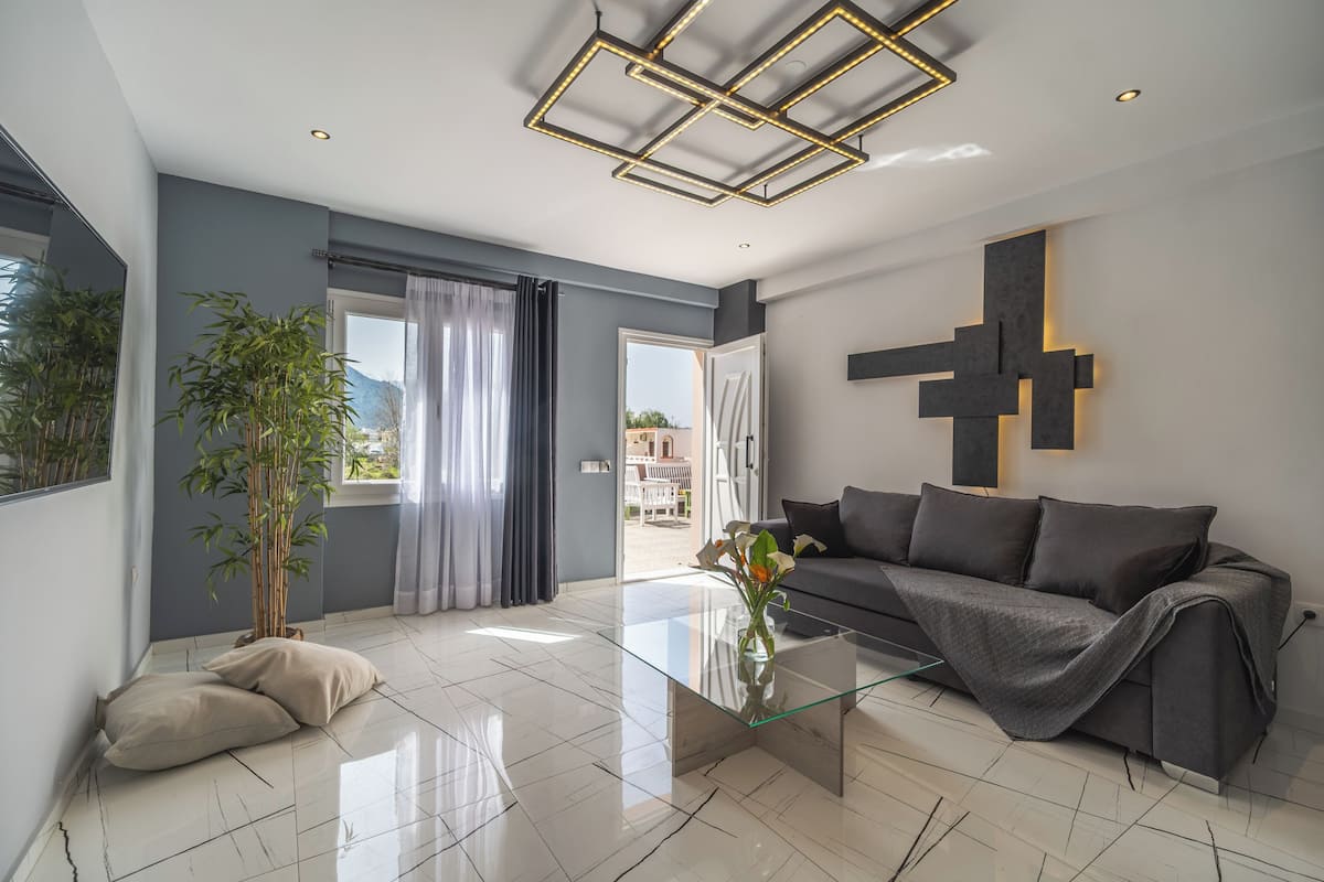A modern living room with a glass coffee table, grey sofa, and a large window with curtains.