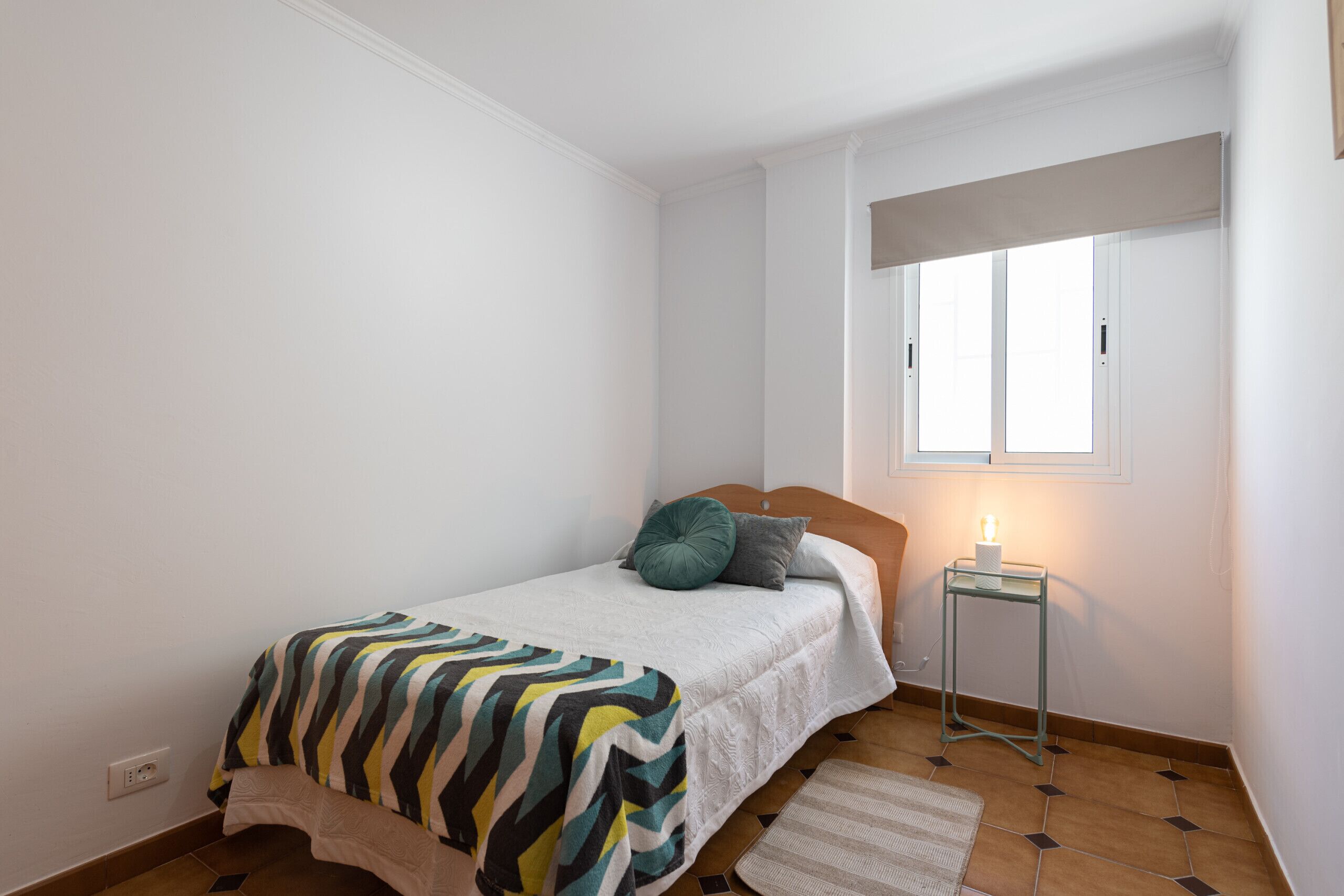 A bedroom with a bed, a bedside table with a lamp, a window, and a rug.
