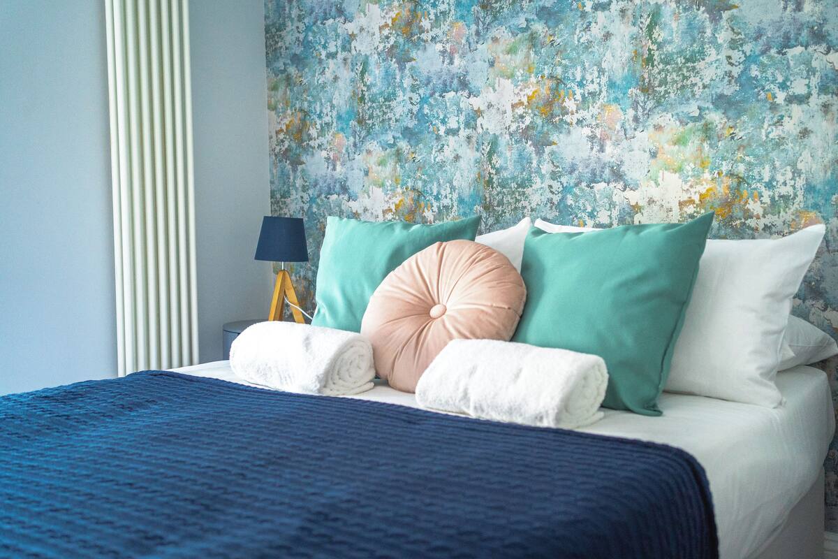 A neatly made bed with a blue blanket, multiple pillows in various colors, and a wall with a distressed paint finish.