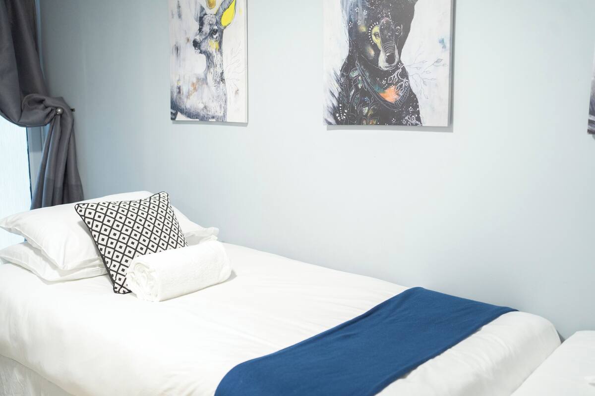 A hotel room with a neatly made bed, two abstract paintings on the wall, and a window with curtains.