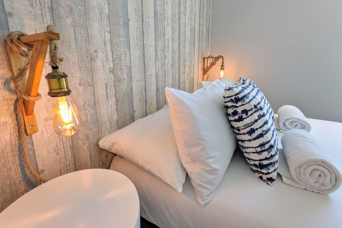 A neatly made bed with white linens and a striped pillow, a round white stool, and a wall-mounted light fixture.