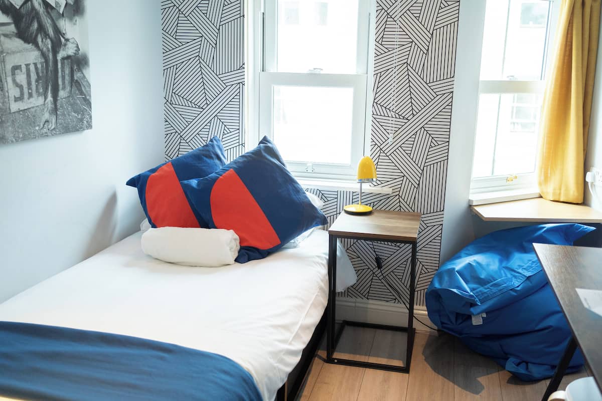 A bedroom with a bed, a desk, and a bean bag chair. There is a window with curtains and a wall with a geometric pattern.