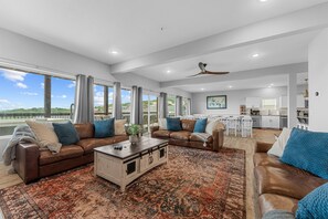 A spacious living area with a large window, a ceiling fan, and a kitchen in the background.
