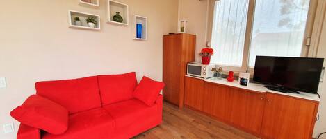 A red sofa in a room with a wooden cabinet, a television, and a window with curtains.