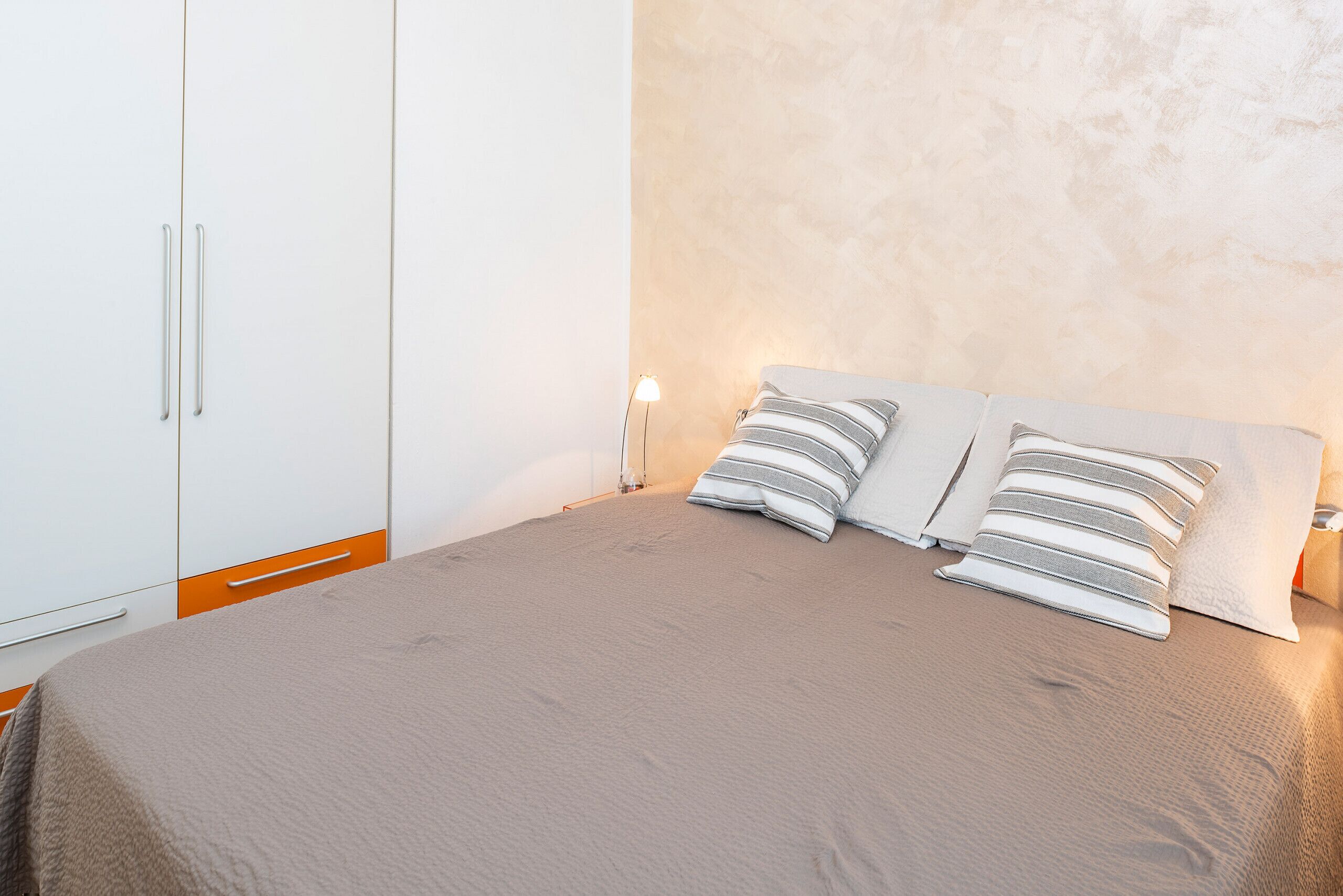 Een slaapkamer met een bed, twee kussens, een nachtkastje, een lamp en een kledingkast.