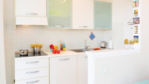 Une cuisine moderne avec des meubles blancs, une cuisinière en acier inoxydable et un évier. On y trouve des légumes colorés sur le plan de travail et une bouilloire.