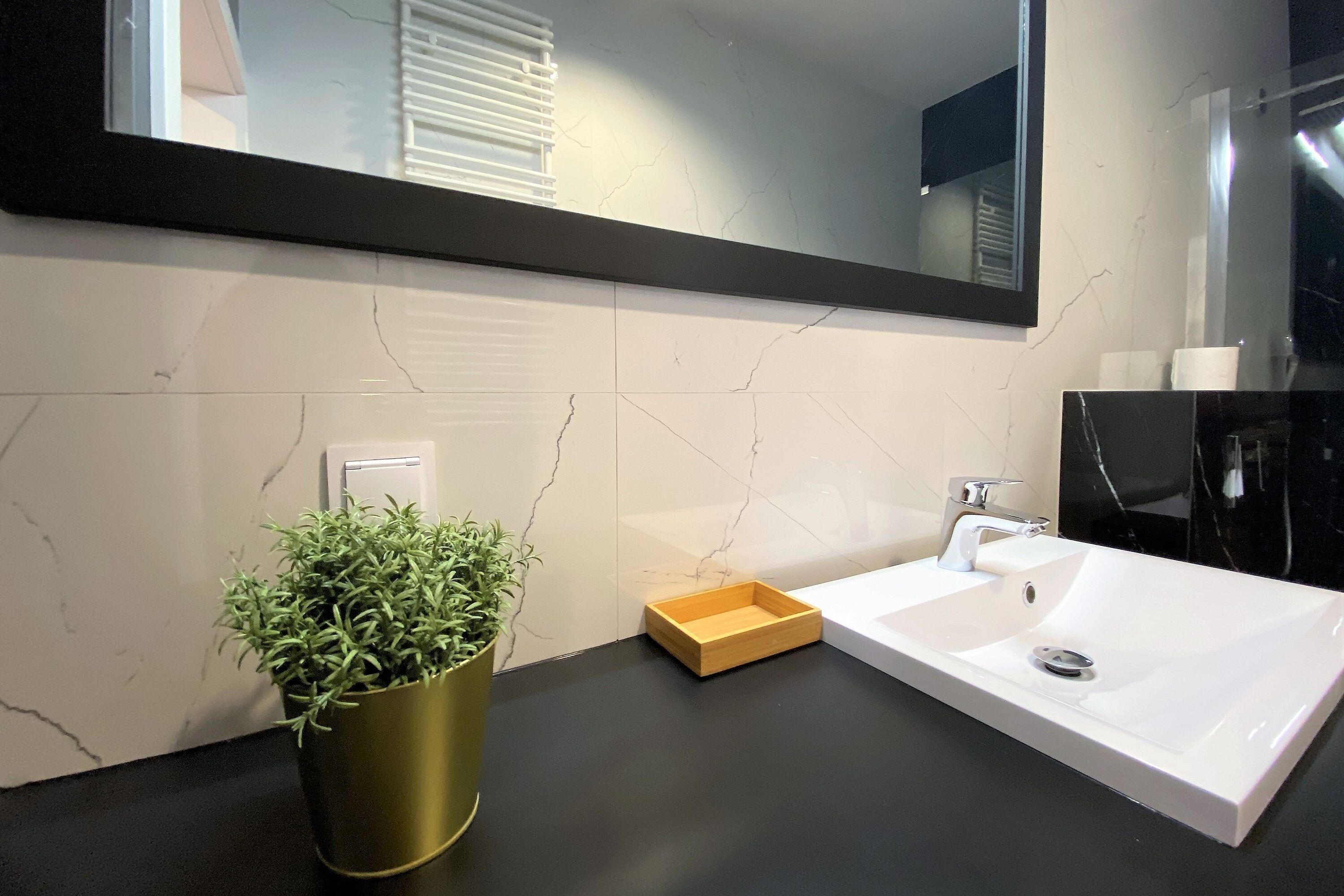 Un baño moderno con un espejo grande, un lavabo blanco, una planta en maceta y una bandeja de madera.
