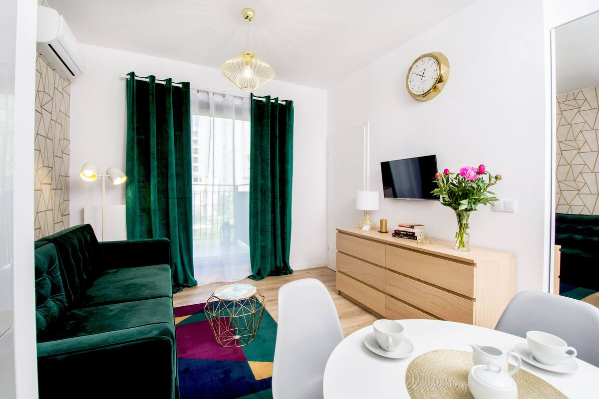 A modern living room with a green sofa, a white dining table, and a TV on the wall.