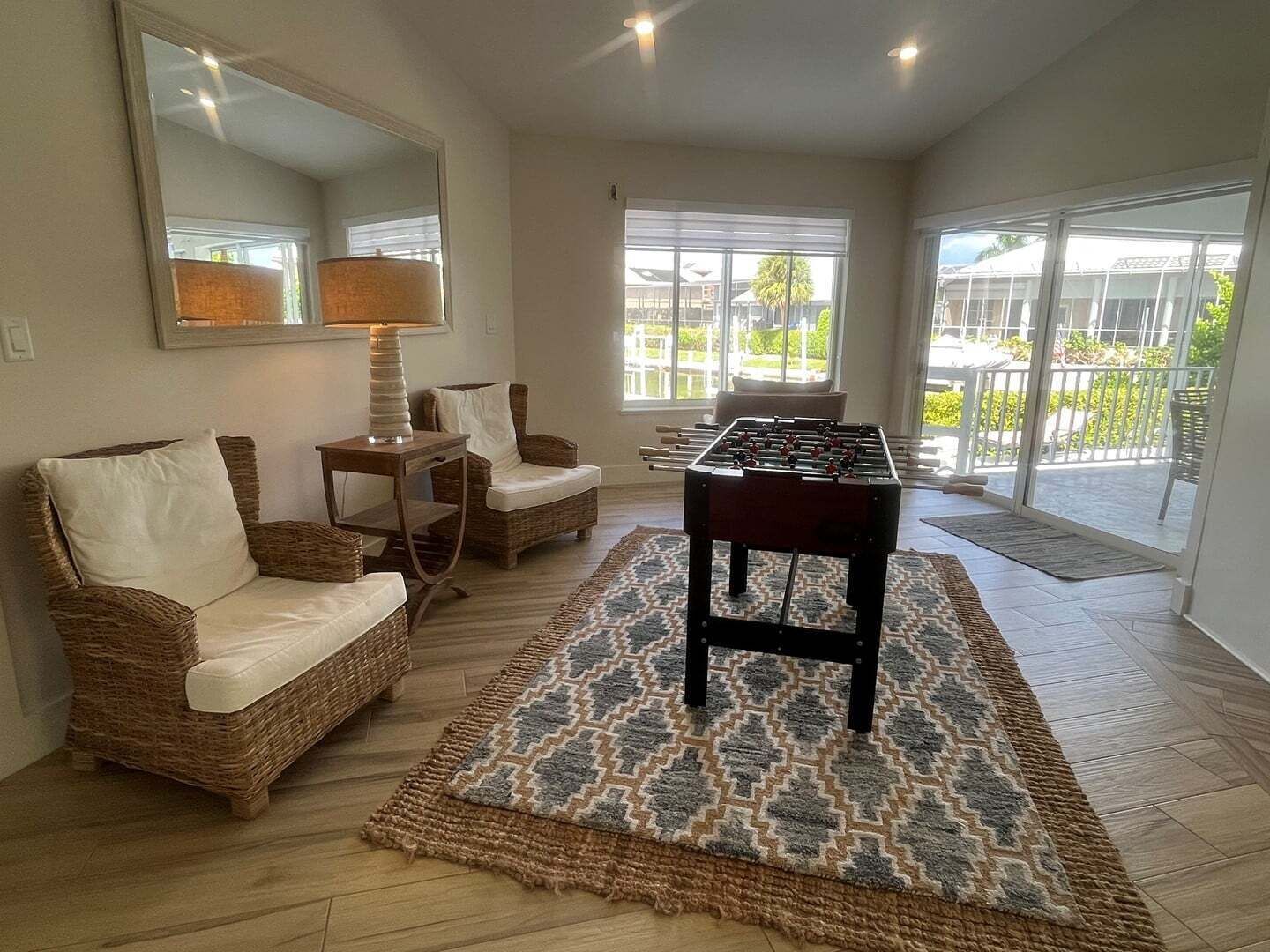 Une pièce avec une table de baby-foot, des meubles en osier, un tapis à motifs et de grandes fenêtres.