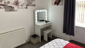 A bedroom with a white vanity, a mirror with lights, a radiator, a small stool, a red blanket, and artwork on the walls.