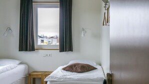 A room with two single beds, a wooden table, a window with curtains, and a view of the outside.
