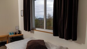 A hotel room with a bed, a desk with a chair, a window with curtains, and a view of trees and hills outside. - Vogahraun 4 (Myvatn)