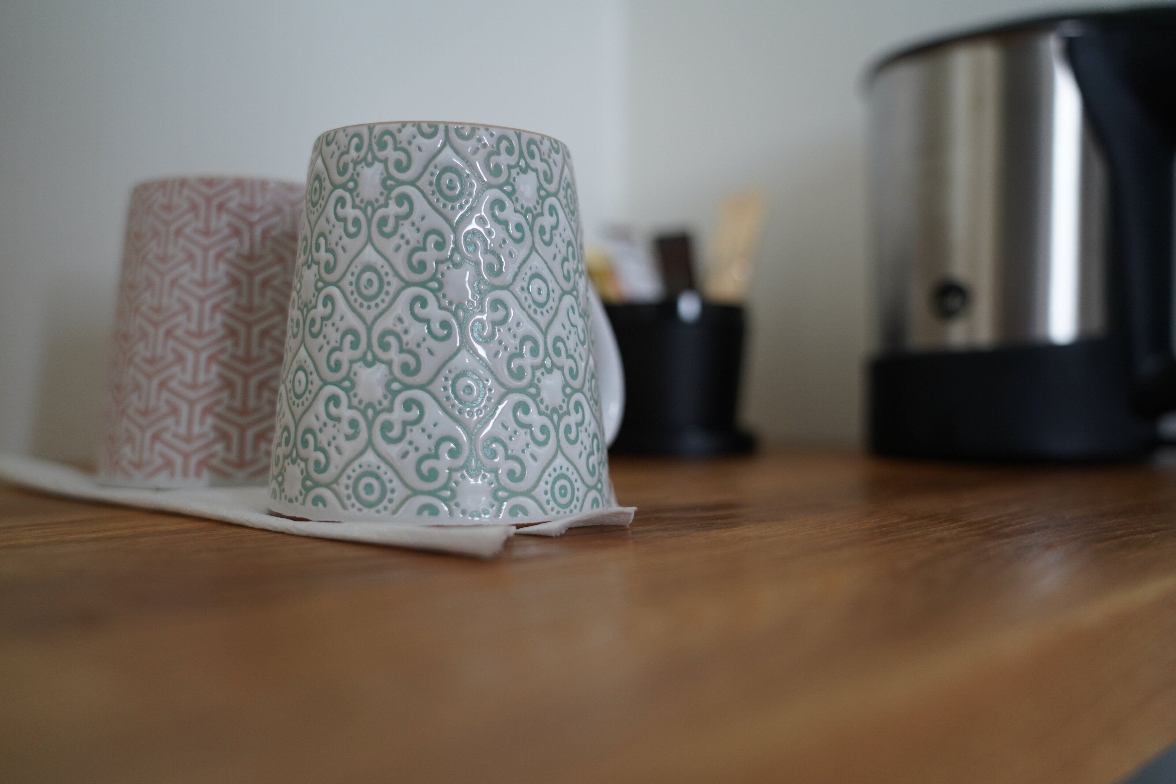Deux tasses à motifs posées sur une surface en bois, avec un appareil en acier inoxydable en arrière-plan.