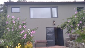 Een gebouw met een grijze gevel, een zwarte deur en een raam. Voor het gebouw staan een bankje en een lantaarnpaal.