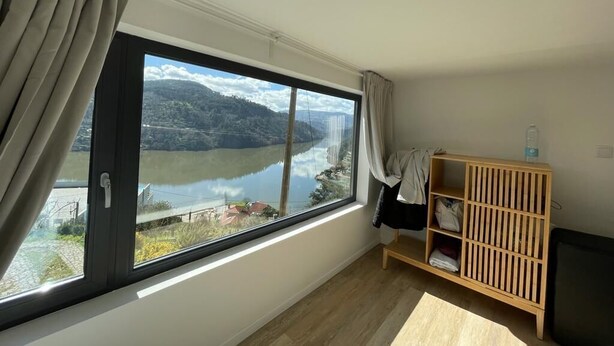 Een kamer met een groot raam dat uitzicht geeft op een meer en bergen. Er is een houten plank met een waterfles erop.