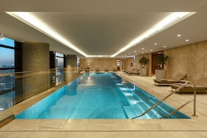 A modern indoor swimming pool with a glass barrier, lounge chairs, and a city view.