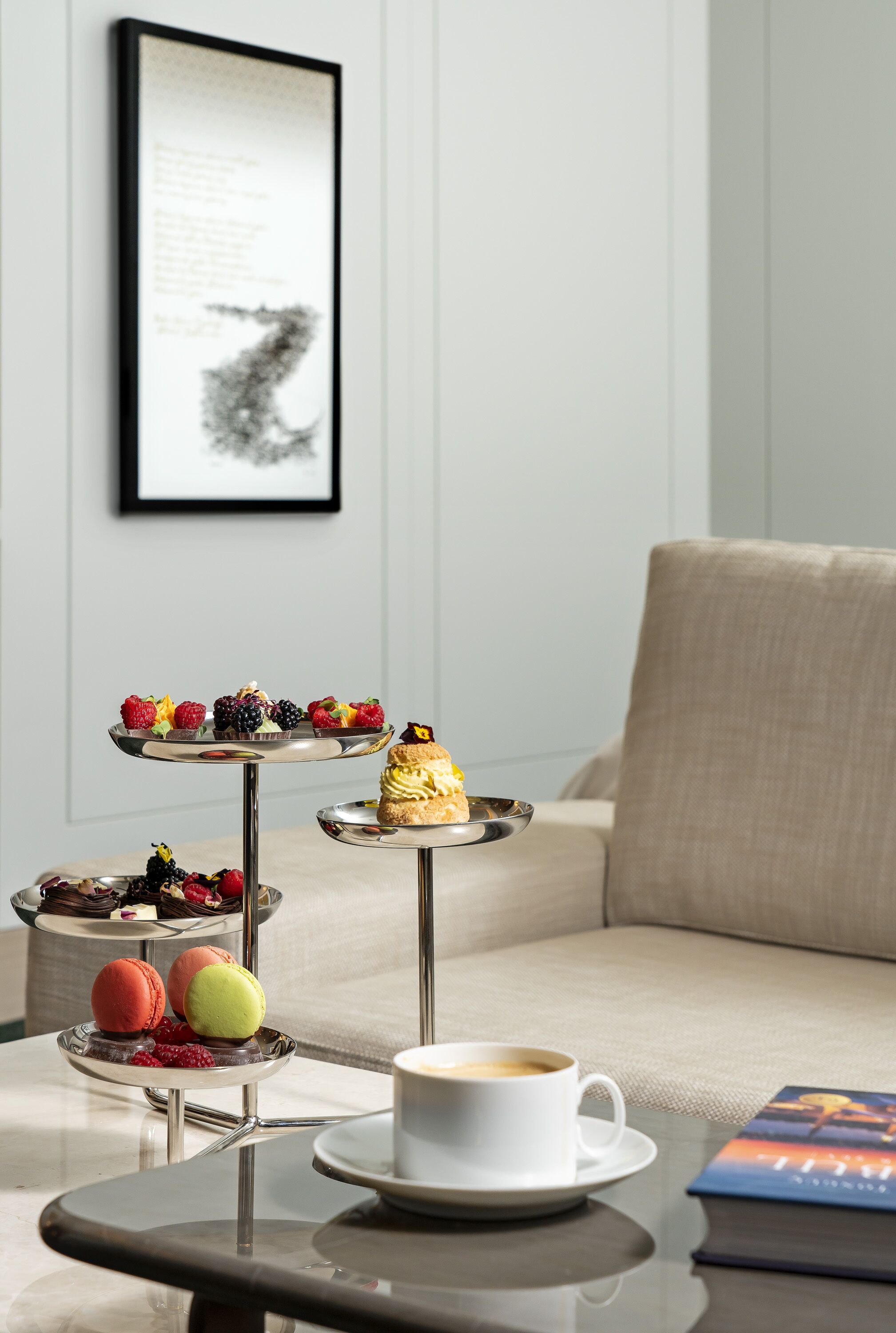 A three-tiered dessert stand with various pastries and fruits, a white cup of coffee, and a book on a table in a modern room with a beige sofa.