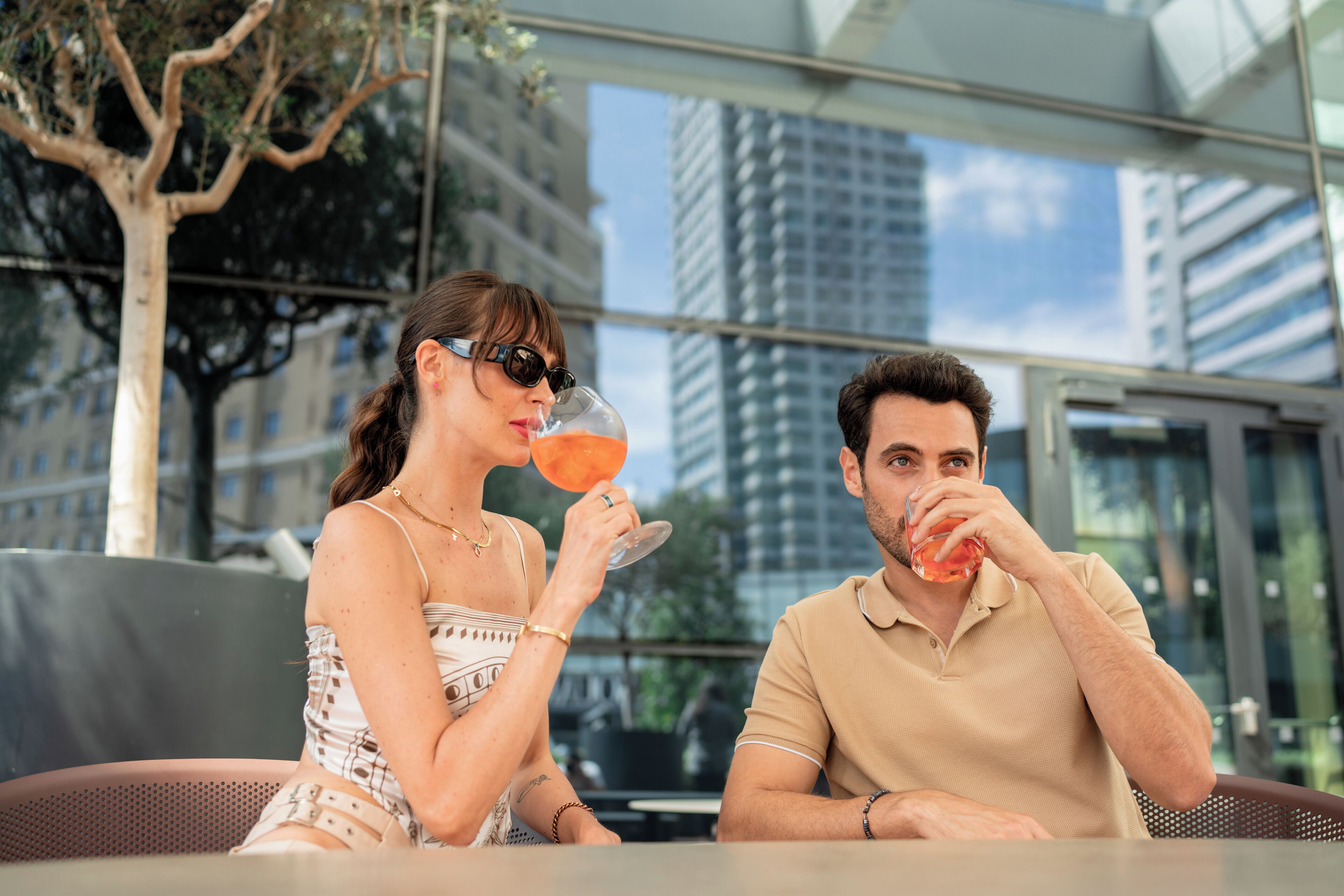 Deux personnes sont assises à une table en plein air, avec vue sur la ville, et l’une d’elles tient un verre à la main.