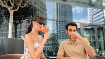 Deux personnes sont assises à une table en plein air, avec vue sur la ville, et l’une d’elles tient un verre à la main.