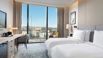 Habitación de hotel con dos camas, un escritorio, una silla, una lámpara y un ventanal con vista a la ciudad.