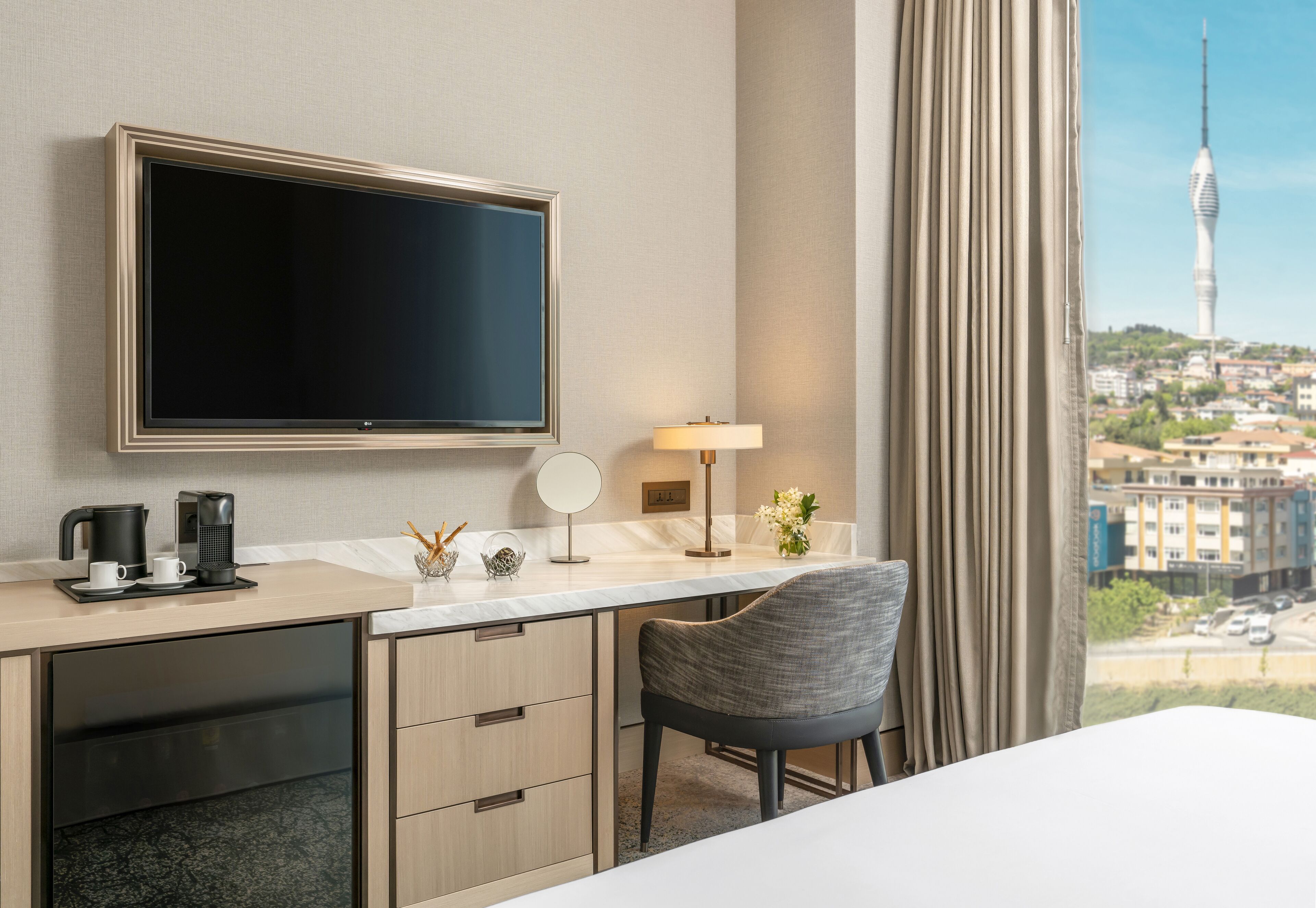 Habitación de hotel con una televisión de pantalla plana grande, un escritorio con cafetera, una lámpara y una silla. La habitación tiene vista a un paisaje urbano con una torre alta.